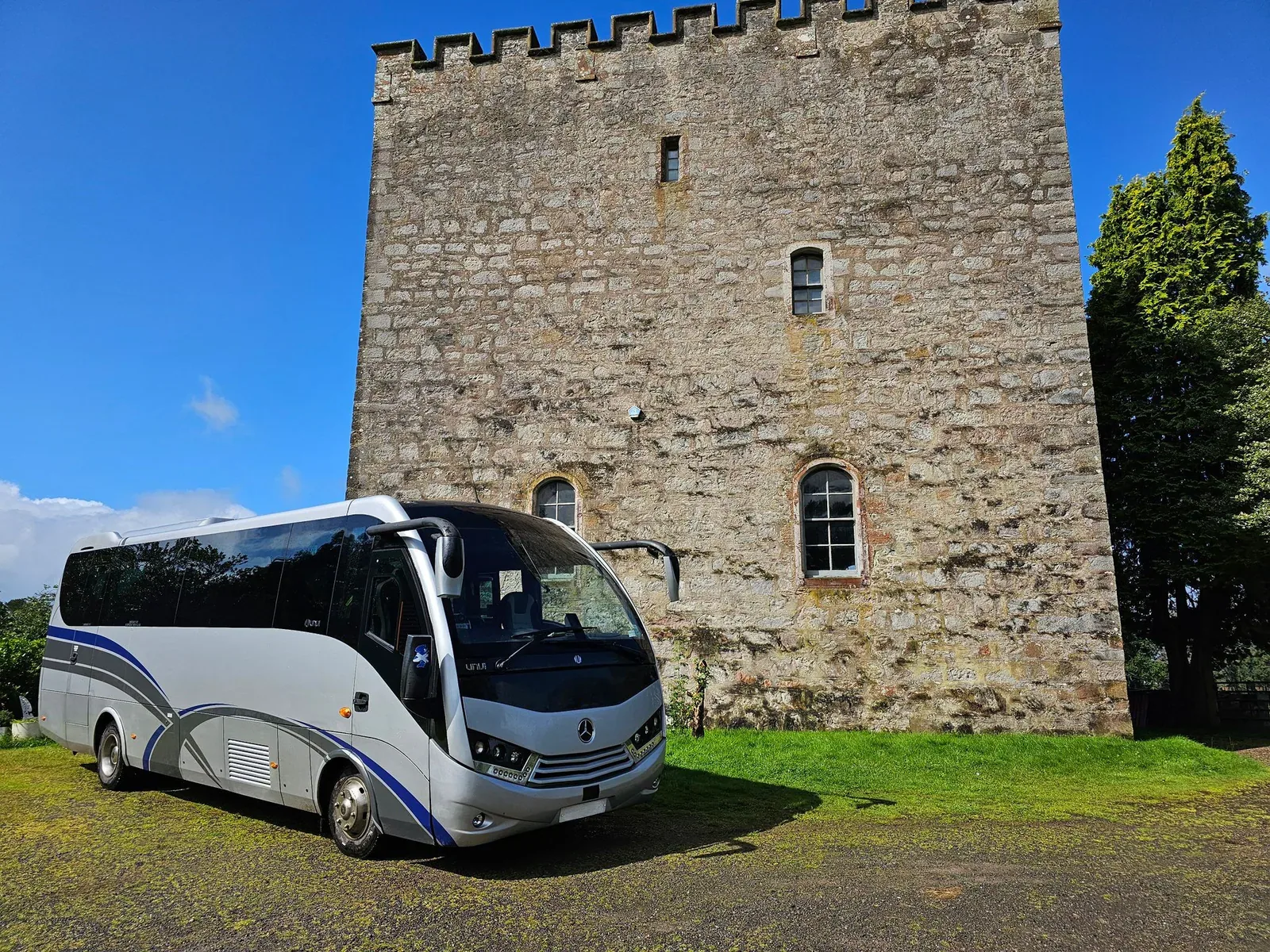 Silver coach with blue and grey side graphics beside a stone tower.